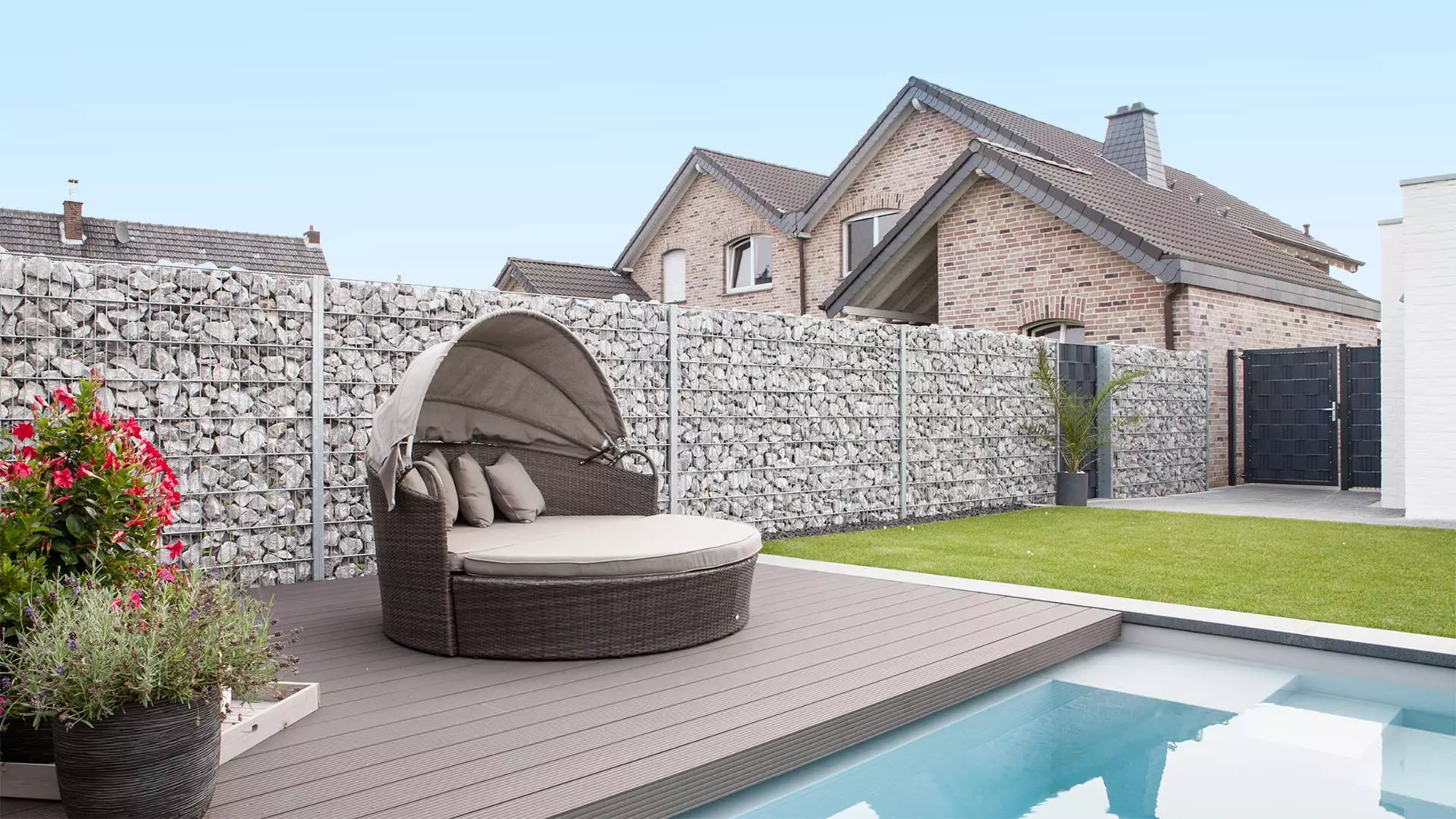 Runder, gepolsterter Outdoor-Liegestuhl mit Überdachung auf Holzterrasse neben Pool und Rasenfläche vor Gabionenmauer