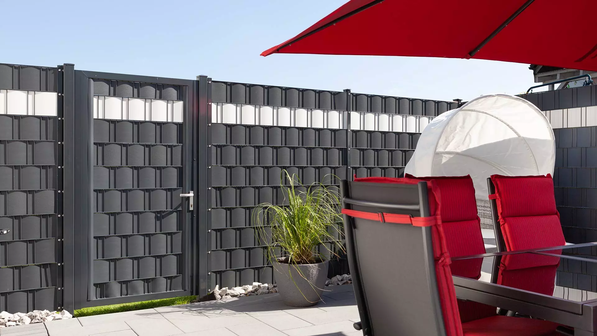 Anthraziter Sichtschutzzaun, davor eine Terrasse mit Sonnenschirm, Gartenstühlen und Tisch
